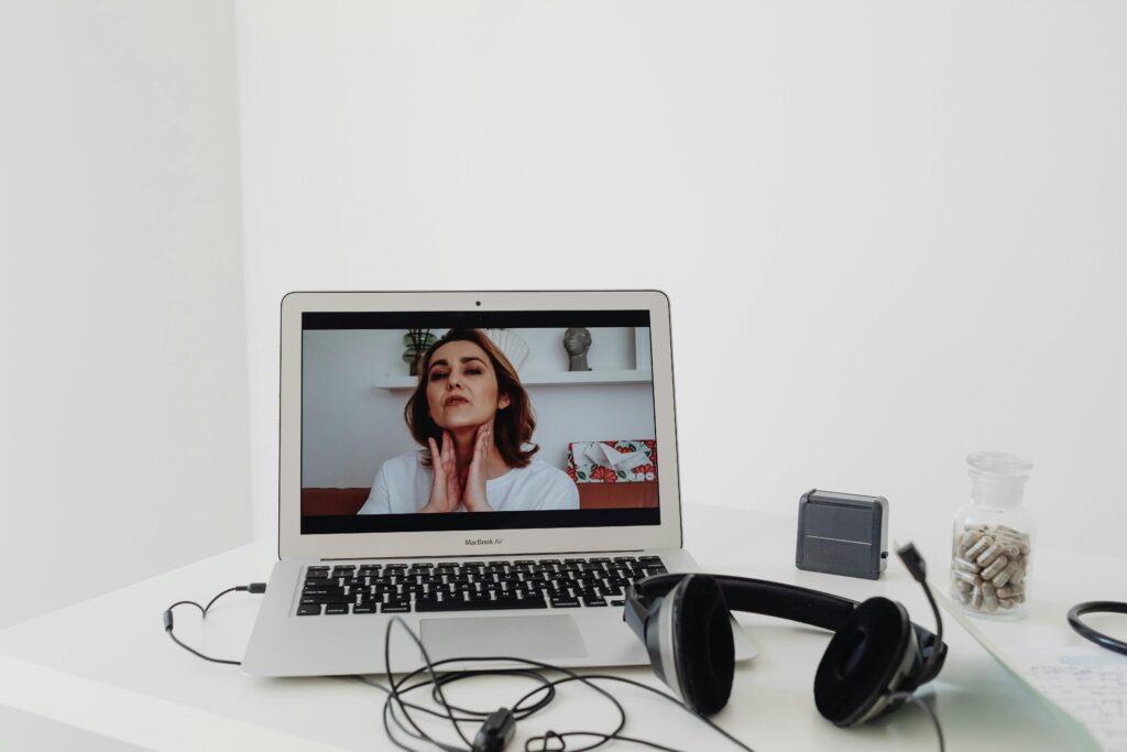 A woman conducting a telemedicine session visible on a laptop screen with headphones nearby.
