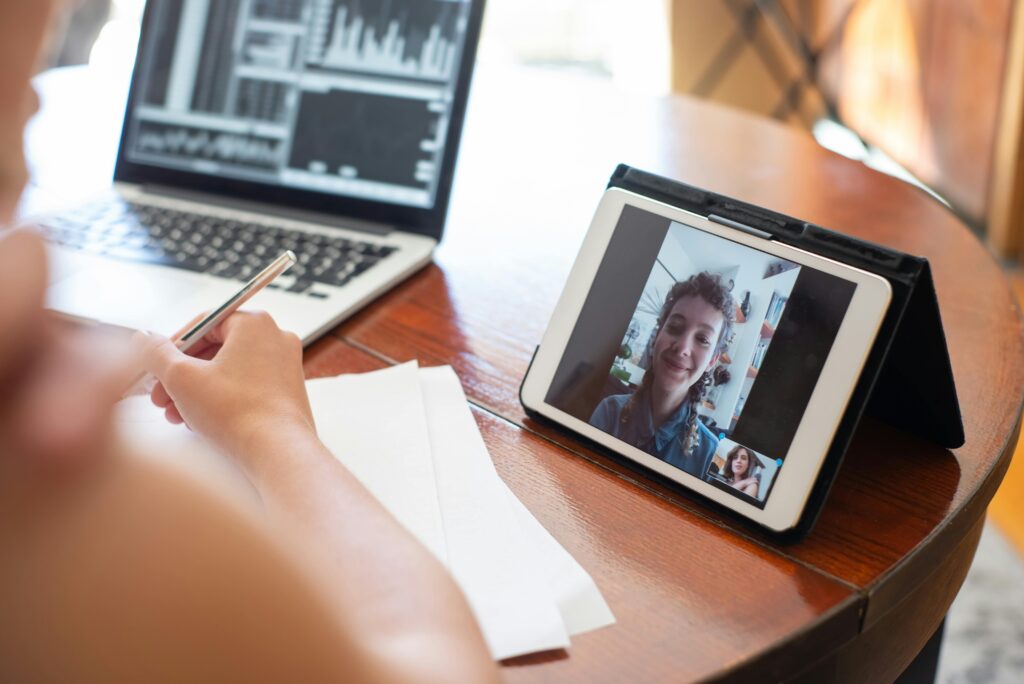 A person on a video call using a tablet, with a laptop displaying data nearby.